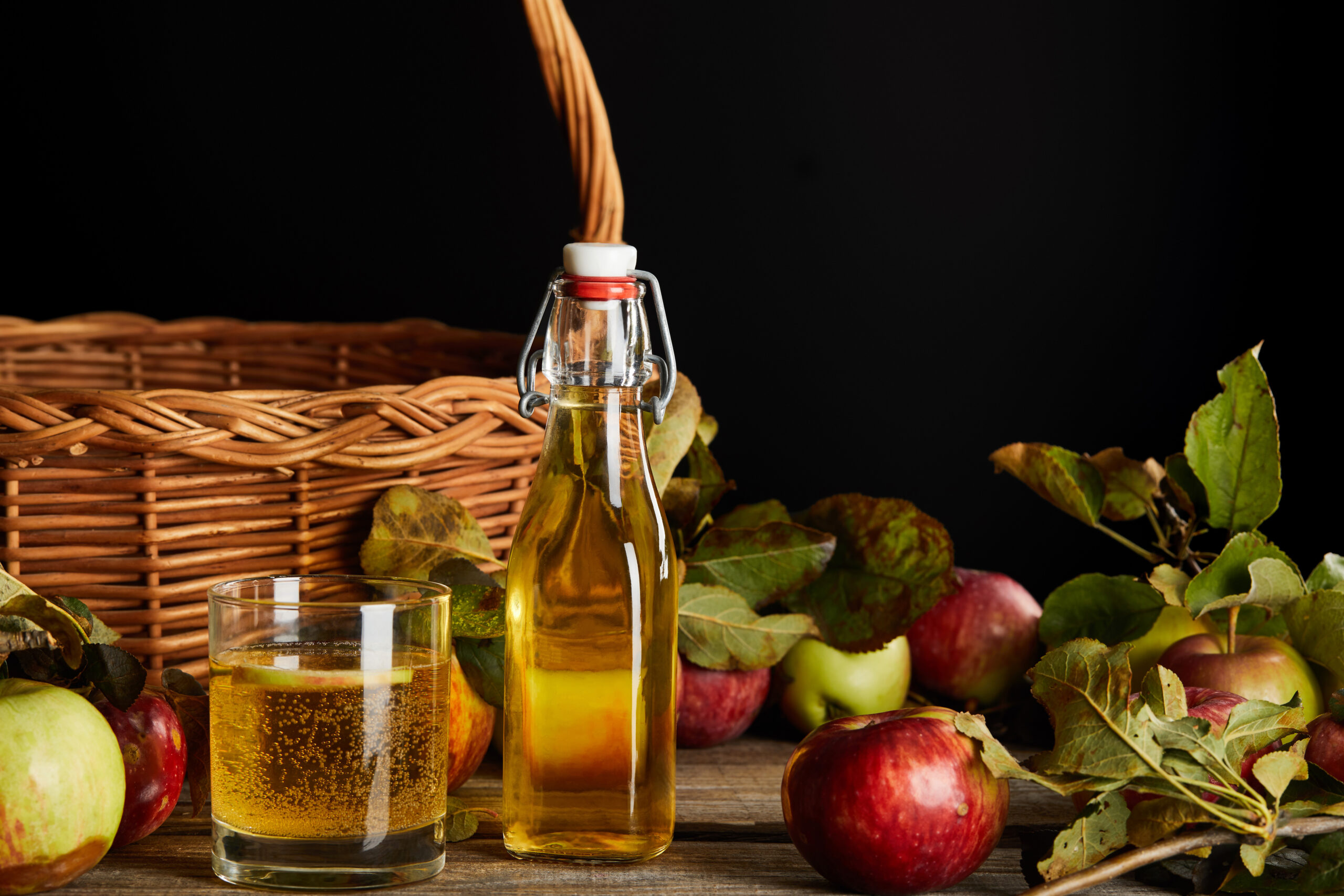 Bottle of apple cider vinegar with a glass of drink, surrounded by fresh apples and leaves, on a wooden surface, highlighting natural haircare benefits.
