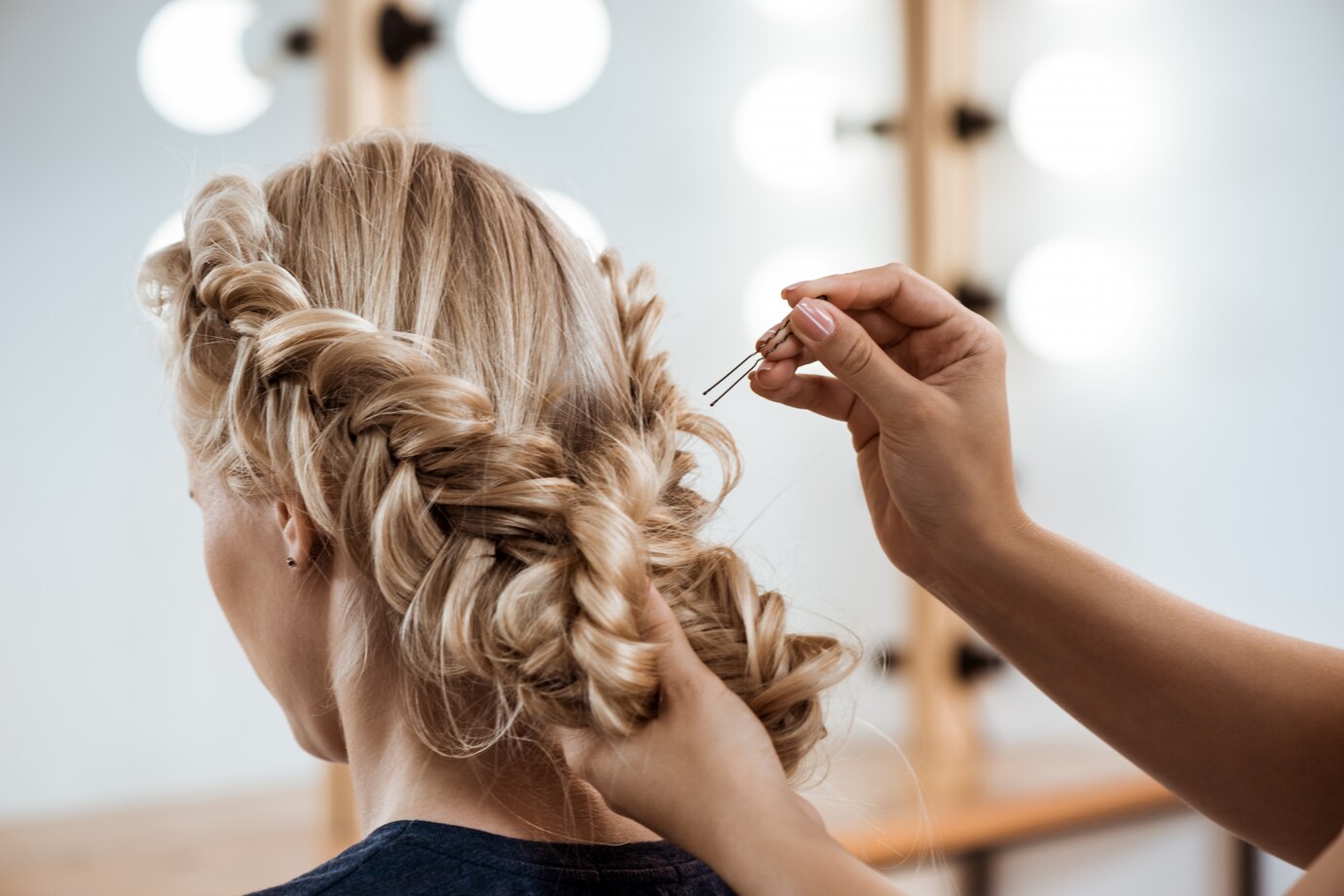 Female hairdresser styling a blonde woman's hair in a beauty salon, showcasing intricate braiding techniques and hairpin application, emphasizing personal care and confidence in hair treatment.