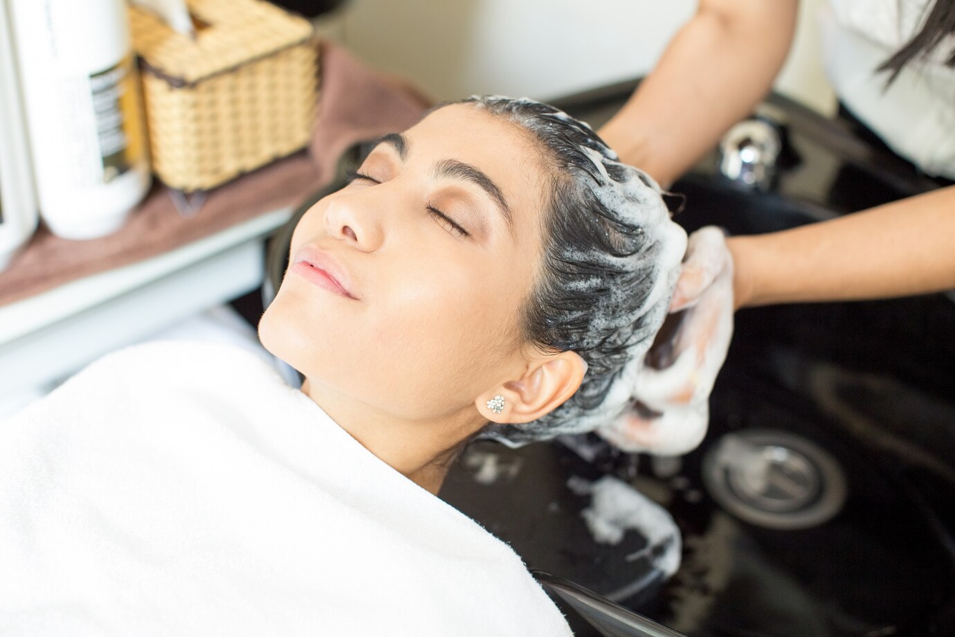 Young woman relaxing in salon chair while receiving a hair wash, with shampoo lather and attentive stylist, emphasizing hair care and self-care journey.